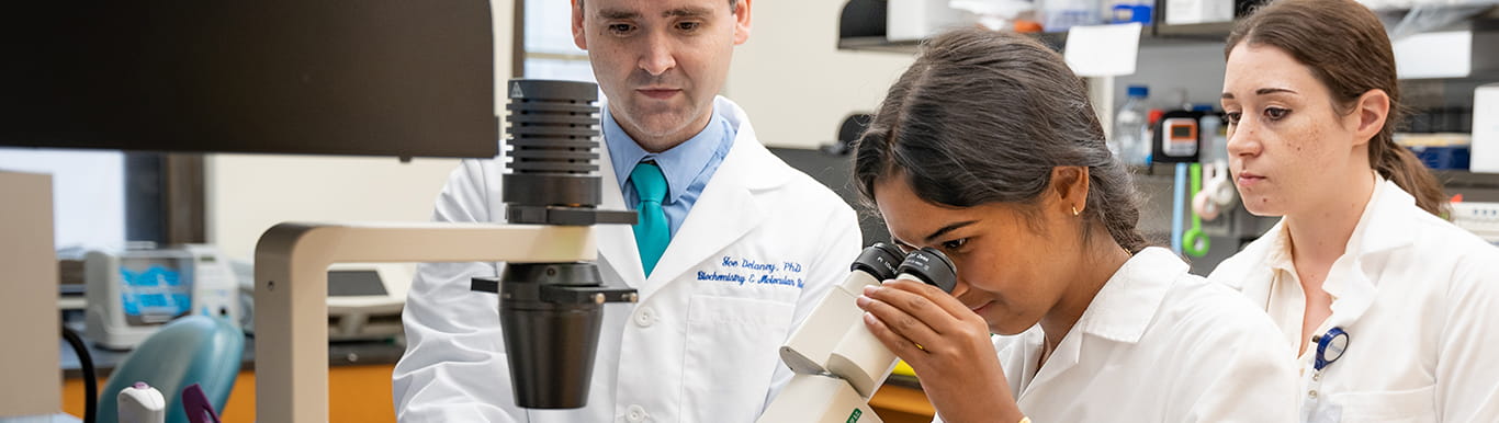 a woman looks into a microscope in a research lab while a man and a woman look on behind her